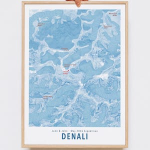May include: A framed topographic map of Denali in shades of blue, featuring contour lines and labeled mountain peaks with elevations in meters. The bottom text reads June & John - May 2026 Expedition and DENALI in bold blue capital letters.