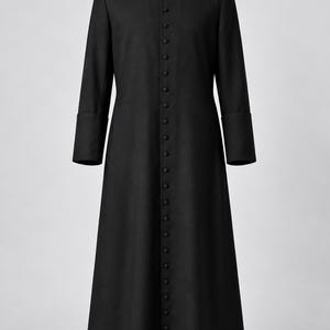 May include: A black cassock featuring a high mandarin collar with a white clerical insert, long sleeves with wide cuffs, and a full-length front button placket with numerous small black buttons extending from the neck to the hem.