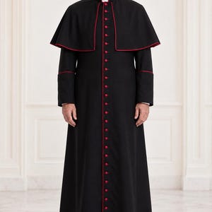 May include: A black cassock featuring a high collar, long sleeves, and a floor-length hem. The garment is accented with red piping along the edges and a vertical row of red buttons down the front, paired with a white clerical collar insert.