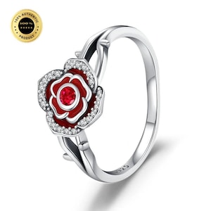 Peut inclure: Une bague en argent avec un motif de rose rouge centrale ornée d'une pierre circulaire rouge vif en son cœur. Les pétales de la rose sont soulignés d'argent et agrémentés de petites pierres claires sur les bords. L'anneau imite une tige épineuse.