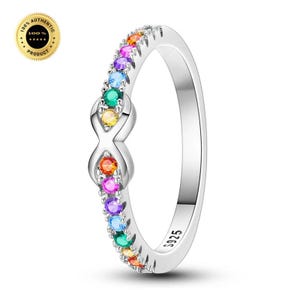 Pode incluir: Um anel de tom prateado com um elemento central em forma de infinito e uma série de pequenas pedras preciosas redondas e multicoloridas cravadas ao longo da banda. O anel de metal tem gravado o texto S925, indicando prata de lei.