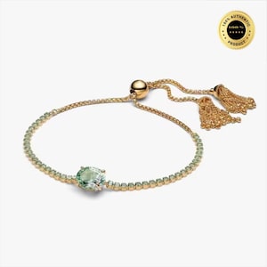 Op de afbeelding: Een verstelbare goudkleurige armband met een rij kleine lichtgroene steentjes en een grotere ovale lichtgroene steen in het midden. De armband heeft een schuifsluiting met twee bungelende goudkleurige kwastjes aan de uiteinden.