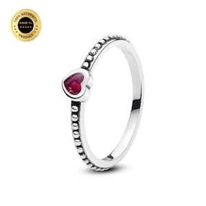 May include: A silver ring featuring a small, heart-shaped red gemstone set at the top. The band is constructed with a series of raised, rounded beads along its entire circumference, creating a textured appearance. The metal has a polished finish.