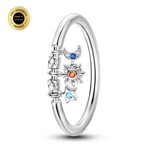 May include: A silver-toned ring featuring a dangling charm element with a crescent moon, a sun, and a star. The moon is set with a blue stone, the sun with an orange stone, and the star with a light blue stone, all accented by clear crystals.