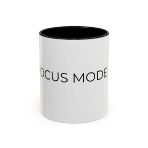 May include: A white ceramic mug with a black interior and rim. The exterior features the text FOCUS MODE printed in a simple, black, sans-serif font. The mug has a cylindrical shape and a smooth, glossy finish, displaying clear, legible lettering.
