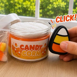 May include: A clear plastic jar filled with orange and yellow slime topped with white foam, featuring a label that reads CANDY CORN. Beside it is a small plastic bag containing candy corn pieces and a black-rimmed candy corn shaped charm.