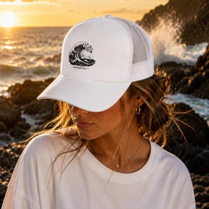 May include: A white trucker cap featuring a black graphic print of a large ocean wave and a mountain peak, with the text "MORBEAL" printed underneath the illustration. The cap has a solid front panel and a breathable mesh back panel.