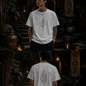 Può includere: Una t-shirt bianca a maniche corte con una grafica nera di un serpente avvolto sul petto con il testo MORBEAL. Il retro della maglietta mostra un'illustrazione più grande di un serpente nero affiancata da caratteri giapponesi verticali.