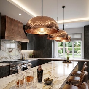 May include: Three dome-shaped pendant lights with a hammered copper finish suspended from black cords. Each light features a warm glowing bulb inside the metallic shade, providing downward illumination over a marble kitchen island surface.