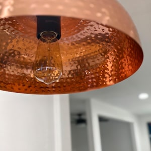 May include: Close-up of a copper pendant light fixture with a hammered texture. The light bulb is visible, suspended from a dark brown socket. The fixture is a warm, metallic color, reflecting the light.