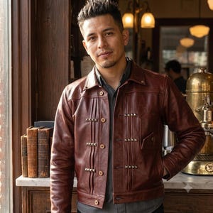 May include: A brown leather jacket featuring a pointed collar, long sleeves, and a button-down front. The front panel displays vertical stitched pleats accented with rows of small, square metallic studs. A single chest pocket is visible.