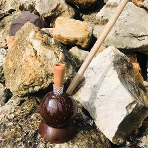 May include: A handcrafted smoking pipe featuring a polished dark brown coconut shell base, a light brown clay bowl attached with a clear mesh connector, and a long, slender wooden stem extending diagonally from the side of the coconut shell.