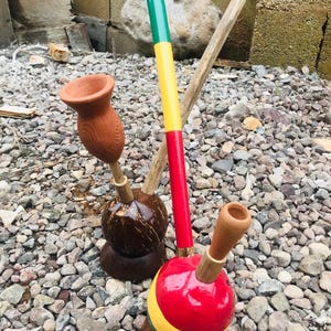May include: Two smoking pipes made from coconut shells and clay. One pipe features a natural brown coconut base with a terracotta bowl. The other pipe has a red, yellow, and green painted coconut base with a long, multi-colored wooden stem.