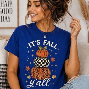 Peut inclure: Un t-shirt bleu roi à manches courtes avec une impression graphique de trois citrouilles empilées. La citrouille du haut est orange, celle du milieu a un motif à damier noir et blanc, et celle du bas a un imprimé léopard. Texte : IT'S FALL y'all.