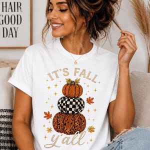 Peut inclure: Un t-shirt blanc à manches courtes avec un motif de trois citrouilles empilées aux motifs orange, à carreaux noirs et blancs, et léopard. Le texte indique IT'S FALL y'all, entouré de petites étoiles dorées et de feuilles d'automne.