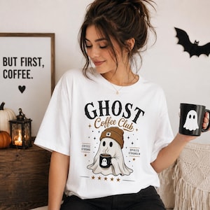 Peut inclure: Un t-shirt blanc avec un motif de fantôme portant un bonnet marron et tenant une tasse de café. Le texte indique "GHOST Coffee Club", "COFFEE STRONG" et "SPIRITS STRONGER". Le design comprend de petites étoiles et une note de cinq étoiles.