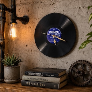 Peut inclure: Une horloge murale fabriquée à partir d'un disque vinyle noir avec une étiquette centrale bleue Philips et des aiguilles dorées. Le disque est de forme circulaire et est fixé sur un mur en béton texturé, affichant le texte Philips 33 1/3 RPM.