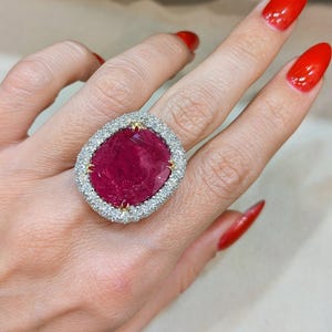 Pode incluir: Um anel grande de formato oval com uma pedra preciosa vermelha profunda e proeminente no centro, rodeada por um halo de numerosas pedras brancas pequenas e brilhantes, fixadas numa armação metálica com quatro garras douradas de suporte.
