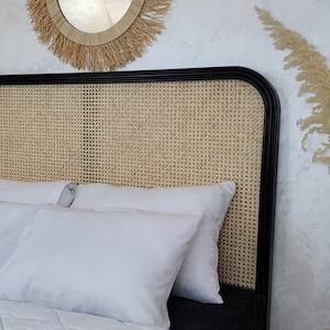 May include: A black bed headboard featuring a natural woven cane panel insert. The bed is dressed with white pillows and a white quilted comforter. Two black vases containing tall dried pampas grass plumes are placed on either side of the bed.