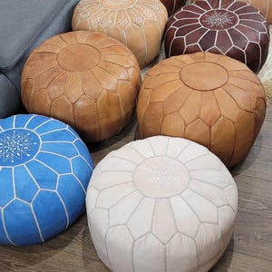 May include: A collection of eight round, stuffed poufs in various colors including blue, white, tan, and brown. Each pouf features a circular top panel with intricate white stitched embroidery and a segmented design radiating from the center.