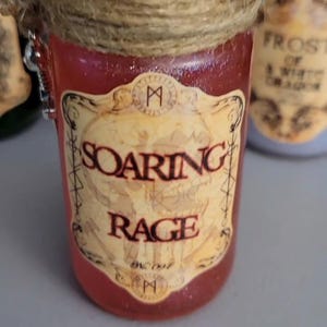 May include: A small, cylindrical jar with a red liquid and a label that reads "SOARING RAGE." The jar is topped with black stones and wrapped with twine. The label has an antique design.