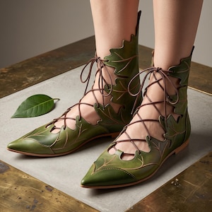 May include: A pair of olive green pointed-toe ankle boots featuring a cutout design shaped like overlapping leaves. The boots are secured with thin brown laces that crisscross up the front and sides, creating a structured, botanical-inspired look.
