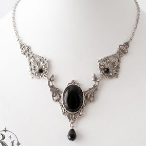 May include: A silver-toned necklace featuring an oval black cabochon centre stone, ornate filigree metalwork, and two smaller black accent stones. A small black teardrop-shaped bead hangs from the bottom of the central oval setting.
