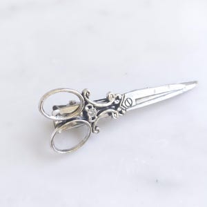 Peut inclure: Une pince à cheveux en métal argenté en forme de paire de ciseaux anciens. L'accessoire présente des détails complexes en volutes le long du manche et des lames, avec une petite pierre claire sertie au centre du motif décoratif.
