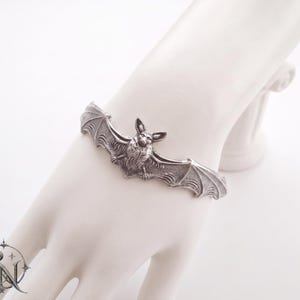 Op de afbeelding: Een zilverkleurige metalen armband met een gedetailleerde, gebeeldhouwde vleermuis met uitgestrekte vleugels. Het vleermuisontwerp is gecentreerd op de band en toont textuurrijke vachtdetails en duidelijk gedefinieerde vleugelmembranen.