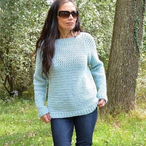 May include: A light blue crochet sweater with long sleeves. The sweater has a textured stitch pattern and a boat neck. The person is wearing blue jeans and sunglasses. The background is a natural outdoor setting with trees and grass.