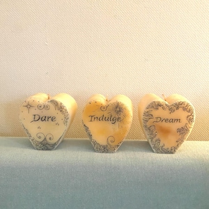 May include: Three heart-shaped cream-coloured candles featuring black ink illustrations and text. The first candle displays the word Dare, the second candle displays the word Indulge, and the third candle displays the word Dream, all with decorative swirls.