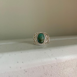 Könnte beinhalten: Ein Silberring mit einem ovalen Türkisstein in der Mitte. Der Stein weist natürliche braune Maserungen auf. Die Ringschiene ist mit filigranen Metallverzierungen und einem Perlenrand um die zentrale Edelsteinfassung gestaltet.