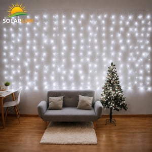 May include: A wall of bright white LED curtain lights hanging behind a gray loveseat with two matching throw pillows. A small decorated Christmas tree stands to the right of the sofa, and a white chair sits at a desk on the left side.