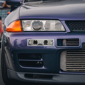 Puede incluir: Una pegatina rectangular para parachoques con fondo gris, un gráfico del monte Fuji y olas estilizadas. La pegatina muestra los caracteres kanji japoneses 神 y 愛 en negro, colocada en el parachoques delantero de un coche deportivo morado.