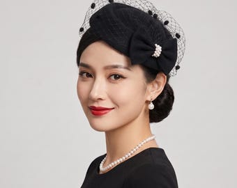 Könnte beinhalten: Ein schwarzer Fascinator-Hut mit einer strukturierten Schleife, die in der Mitte mit einer Gruppe weißer Perlen verziert ist. Das Kopfstück ist mit einem schwarzen Netzschleier akzentuiert, der mit kleinen schwarzen Bommeln besetzt ist.