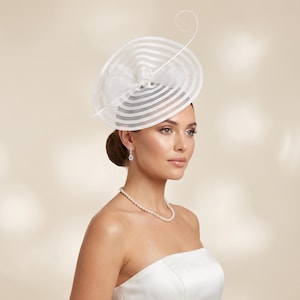 May include: A white fascinator hat featuring a wide, circular brim with concentric sheer stripes and a central fabric bow detail. A thin, curved wire accent extends upwards from the centre. The hat is paired with a simple white pearl necklace.