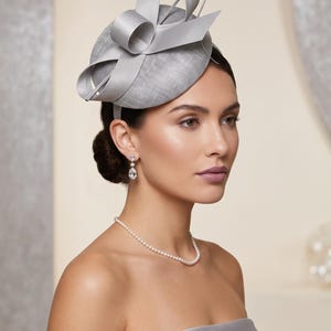 Pode incluir: Um fascinator prateado com uma base circular texturizada, adornado com laços de fita de cetim estruturados e um laço. O acessório é combinado com um colar de pérolas delicado e um par de brincos pendentes transparentes em forma de gota.