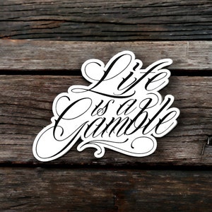 Op de afbeelding: Witte sticker met zwarte, cursieve tekst die "Life is a Gamble" zegt. De sticker is geplaatst op een verweerd houten oppervlak.