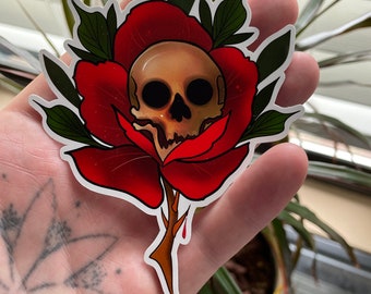 May include: A red rose with a human skull in the centre. The rose is dripping with blood. The sticker is on a hand.