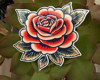 May include: A red rose with green leaves, drawn in a traditional tattoo style. The rose is outlined in black and has a white centre.
