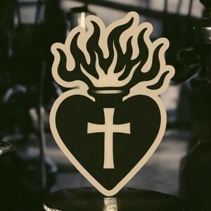May include: A black and white sticker of a heart with flames on top and a cross inside. The heart is outlined in brown.