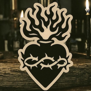 May include: A black and white illustration of a heart with flames on top and a crown of thorns around it. The heart is on a wooden surface.