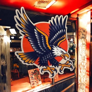 Puede incluir: Un águila negra y blanca con detalles amarillos vuela frente a un sol rojo. El águila es un diseño de tatuaje tradicional.