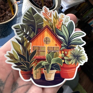Puede incluir: Una pegatina con una ilustración de dibujos animados de una pequeña casa rodeada de plantas en macetas. La casa es amarilla con un techo marrón y ventanas. Las plantas son verdes y marrones con varias formas de hojas. La pegatina está en una mano.