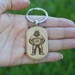 May include: A light-colored wooden keychain with a rounded rectangular shape featuring a laser-engraved illustration of a furry creature standing with hands on hips. The creature wears a t-shirt with an alien head graphic and the text BELIEVE.