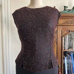 May include: A sleeveless black top featuring an intricate, dense pattern of small, dark red beads covering the entire surface. The garment has a high, rounded neckline and two small vertical slits at the bottom hem on the front side.