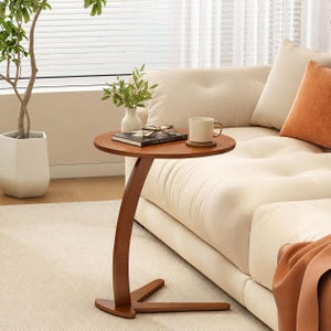 May include: A round, medium-brown wooden side table with a C-shaped base. The table surface holds a white ceramic mug on a small coaster, a black book titled The Kinfolk, and a pair of eyeglasses. The table features a curved support leg.