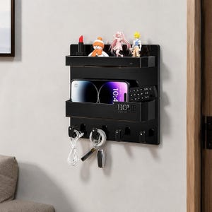 May include: A black wall-mounted organizer featuring two horizontal shelves and five hooks at the bottom. The top shelf holds small figurines and a lipstick, while the middle shelf contains a smartphone and remote. The text HOME is printed on the front.