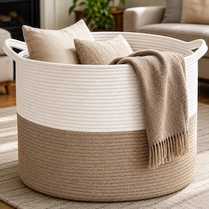 May include: A large, round, two-toned storage basket made of coiled rope, featuring a white upper section and a tan lower section. The basket has integrated handles on both sides and contains two beige throw pillows and a tan fringed throw blanket.