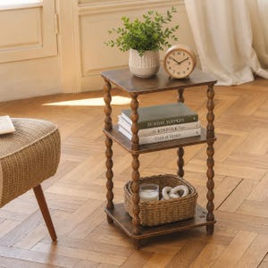 May include: A three-tiered wooden side table with turned spindle legs. The top shelf holds a potted green plant and a round analog clock. The middle shelf contains three stacked books titled Sunday Suppers, The Kinfolk Table, and This Is Home.